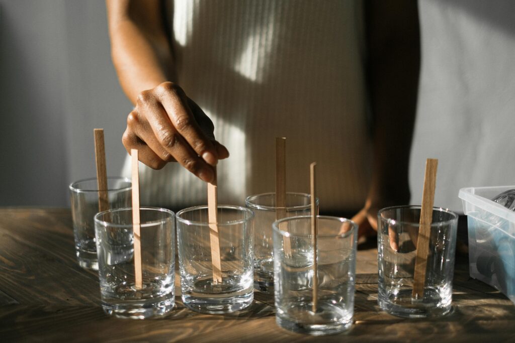 Unrecognizable African American female securing wooden wicks in glass molds while making aroma candles at wooden table in light studio